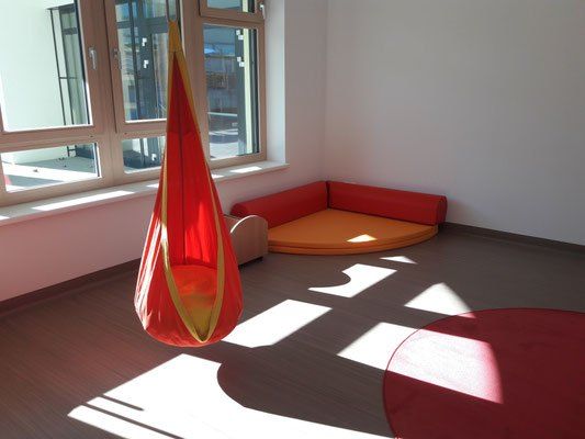 Ein heller Raum mit weißen Wänden und einem Holzboden. Es gibt einen roten Hängesessel und eine rote gepolsterte Couch. Sonnenlicht strömt durch das Fenster.