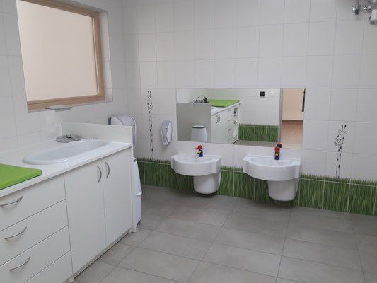 Ein Badezimmer mit weißen Fliesen und Waschbecken, mit einem großen Spiegel, der einen Schreibtisch und ein Fenster reflektiert. Die Wand hat einen grünen Rand und einen Giraffensticker.