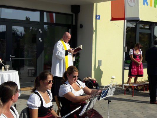 Ein Priester liest draußen vor einem Gebäude ein Buch. Zwei Frauen sitzen und spielen Musikinstrumente. Eine Frau steht im Hintergrund.