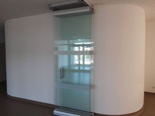 Ein leerer Raum mit einer weißen geschwungenen Wand und einer Glasschiebetür. Die Tür ist geschlossen, und die Reflexion der Außenseite ist auf dem Glas sichtbar.