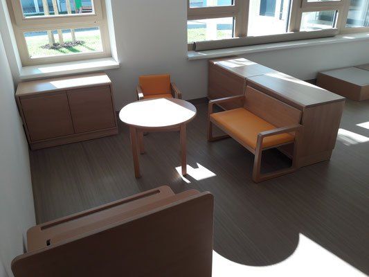 Ein Zimmer mit Holzmöbeln, zwei Fenstern und einem Tisch in der Mitte. Der Raum ist hell, mit Sonnenlicht, das durch die Fenster kommt.