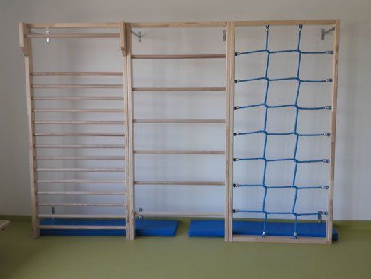 Ein Indoor-Fitnessstudio mit Holzklettergestellen und einem blauen Netz an einer weißen Wand. Blaue Matten auf dem Boden.