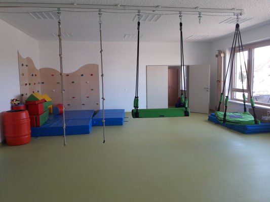 Ein Indoor-Spielplatz mit Schaukeln, Matten und Kletterwänden. Der Raum hat eine weiße Wand und ein großes Fenster mit Blick auf ein Auto.
