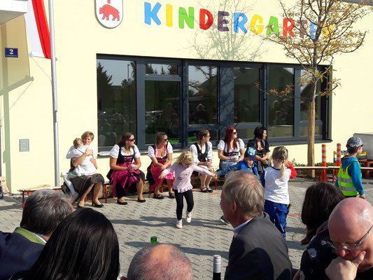 Eine Gruppe von Menschen versammelt sich vor einem Kindergartengebäude. Einige sitzen, andere stehen und ein Kind tanzt. In der Nähe befinden sich eine Fahne und ein Baum.