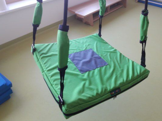 Eine grüne Hängeschaukel mit einem blauen Quadrat in der Mitte hängt von einer weißen Decke. Hinter ihr steht eine Holzbank.
