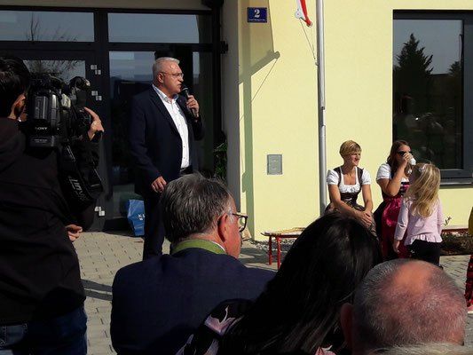 Ein Mann in formaler Kleidung spricht in ein Mikrofon und wendet sich an eine Menschenmenge. Hinter ihm hält eine Frau in traditioneller Kleidung eine Tasse. Die Veranstaltung findet vor einem Gebäude mit Glasfenstern statt.