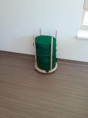 Ein Stapel grüner runder Kissen auf einem hölzernen Rollwagen in einer Ecke eines Raumes mit Holzboden und weißer Wand.