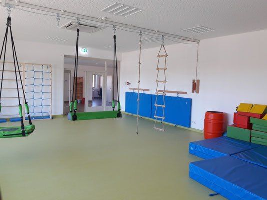 Ein Indoor-Spielzimmer mit zwei hängenden Schaukeln, einer Leiter und blauen Matten. Auf der rechten Seite befinden sich verschiedene farbige Spielzeuge und ein roter Fass.