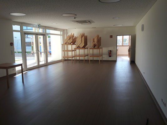 Ein leerer Raum mit Holzboden, Glastüren und einem Feuerlöscher. Es gibt Holzregale mit gestapelten Stühlen.