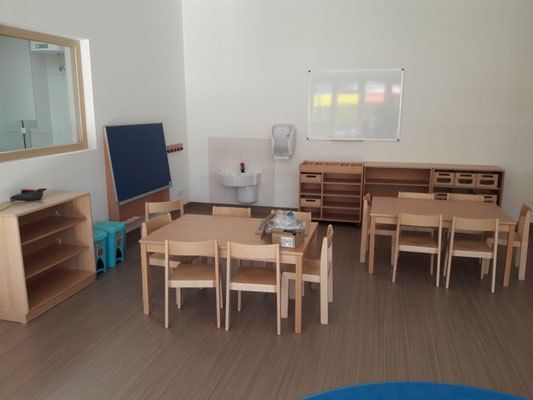 Ein sauberes Klassenzimmer mit Holztischen und Stühlen, Regalen, einem Waschbecken, einer Tafel, einem blauen Brett und einem Spiegel. Links stehen zwei blaue Behälter.