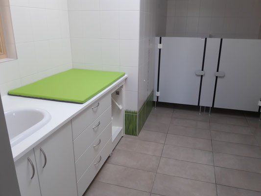 Ein leeres Badezimmer mit weißen Wänden, einem grünen Tisch, einem Waschbecken und drei Kabinen. Der Boden ist gefliest und in der Nähe der Kabinen befindet sich ein grüner und weißer Rand.