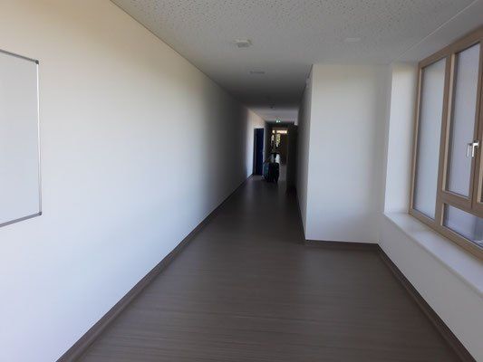 Ein leerer Flur mit weißen Wänden und einem braunen Boden, einer Tafel auf der linken Seite, einer Tür am Ende und einem Fenster auf der rechten Seite.
