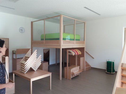Ein Raum mit einem holzfarbenen Etagenbett und einer Frau, die neben einem Tisch mit gestapelten Stühlen steht. Eine Uhr ist an der Wand, und eine Treppe befindet sich rechts.