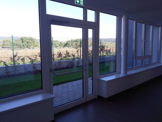 Ein leerer Raum mit Glaswänden und Türen, mit Blick auf ein Grasfeld mit Bäumen und einem Zaun.