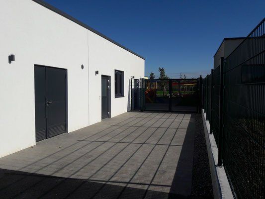 Ein modernes, weißes Gebäude mit zwei Türen, einem gepflasterten Gehweg und einem schwarzen Metallzaun. Im Hintergrund befindet sich ein Spielplatz und ein Rasenfeld.