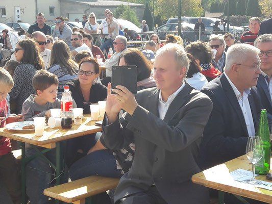 Eine Gruppe von Menschen sitzt draußen an Tischen. Ein Mann hält ein Tablet, andere sitzen mit Tellern und Getränken. Im Hintergrund sind Autos geparkt.