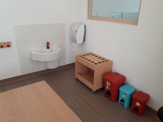 Ein kleines Badezimmer mit Waschbecken, Papierhandtuchspender und Toilette. Es gibt drei Plastikhocker und einen Holztisch. Die Wand ist mit weißen Fliesen verkleidet.