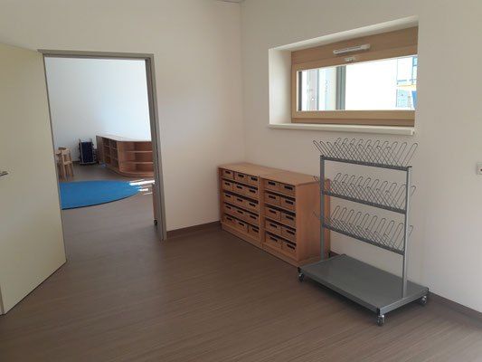 Ein leerer Raum mit Holzboden und weißen Wänden. Ein blauer Teppich liegt auf dem Boden in der Nähe des Fensters. Rechts befindet sich ein Holzschrank mit Schubladen und ein Metallständer.