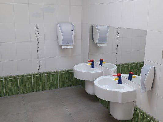 Ein Badezimmer mit zwei Waschbecken, einem Spiegel und Papierhandtuchspendern. Die Wand ist weiß und es gibt einen grünen Grasrand am Boden.
