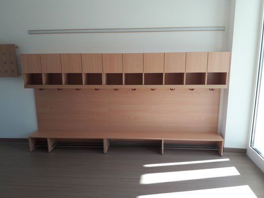Ein leerer Raum mit Holzbänken, Haken und einem Glasfenster. Die Bank hat Fächer für Schuhe und Kleidung, während die Haken zum Aufhängen von Jacken dienen. Der Raum ist durch Sonnenlicht gut beleuchtet.