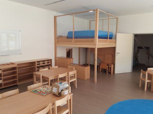 Ein Klassenzimmer mit einem Etagenbett, einer Tafel, einem Schreibtisch und Stühlen. Es gibt einen Holzschrank und Regale mit Aufbewahrungsboxen. Der Raum hat Holzböden und Wände.