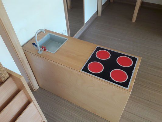 Ein hölzerne Spielküche mit einem Waschbecken und einem schwarzen und roten Herd mit vier Kochplatten. Der Boden ist aus Holz.