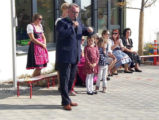 Ein Mann spricht in ein Mikrofon in einem formellen Anzug, während mehrere Frauen und Kinder zuhören. Zwei Frauen in traditioneller Kleidung stehen neben einer Bank.