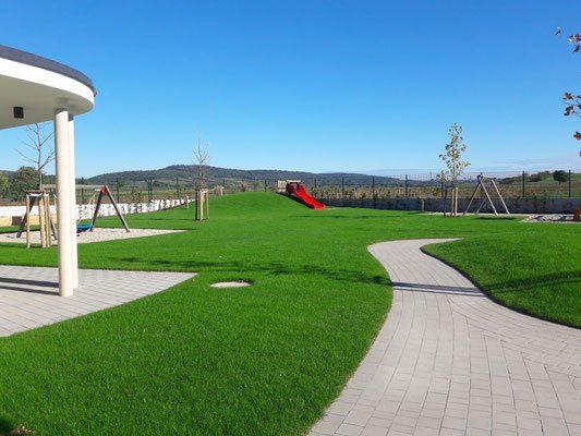Ein Außenspielplatz mit einem roten Rutsch und grünem Gras, umgeben von Bäumen und einem Zaun mit Blick auf die Hügel.
