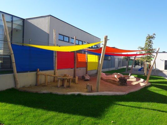 Ein farbenfroher Spielplatz mit Holzkonstruktionen und Sand unter bunten Sonnensegeln. Im Hintergrund befindet sich ein Gebäude mit Glasfenstern.