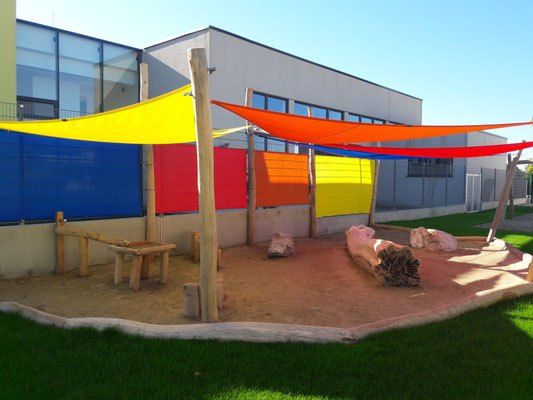 Ein Außenspielplatz mit einer Holzbank, bunten Sonnensegeln und großen Felsen, umgeben von bunten Wänden und Gras.