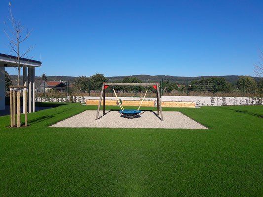Ein Spielplatz im Freien mit einem Schaukelset auf einem gut gepflegten Rasen, einem Haus und entfernten Hügeln unter einem klaren blauen Himmel.