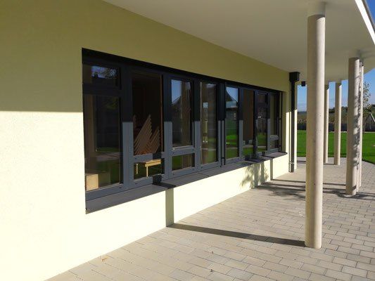 Ein modernes Gebäude mit großen Fenstern und einem gefliesten Boden. Die Fenster haben schwarze Rahmen und reflektieren die Umgebung.