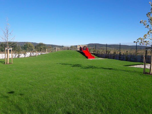 Eine leuchtend rote Rutsche befindet sich auf einem Rasenhügel. Ein Baum und ein Zaun sind in der Nähe. Berge und ein klarer blauer Himmel sind im Hintergrund sichtbar.