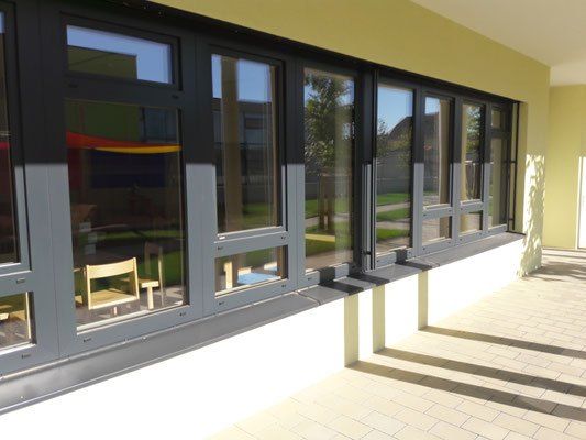 Eine Außenterrasse mit großen Glasfenstern. Die Stühle und der Tisch befinden sich im Raum. Der Schatten des Baumes fällt auf den gefliesten Boden.
