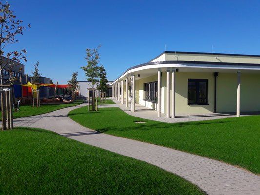 Ein gepflegter Gehweg führt zu einem modernen Gebäude mit kurvenförmigem Dach, umgeben von sattgrünem Rasen und Bäumen unter einem klaren blauen Himmel.