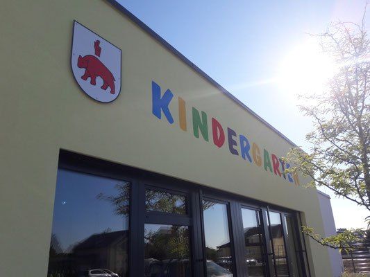 Ein hellgelbes Gebäude mit Glasfenstern und einem roten Bärenlogo. Das Wort 'KINDERGARTEN' ist in bunten Buchstaben an der Wand geschrieben.