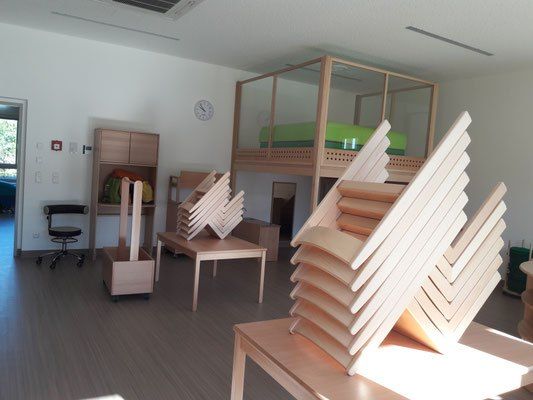 Ein Raum mit Holzmöbeln, einem Etagenbett und einer Uhr an der Wand. Stapel von Holzstühlen sind auf Tischen angeordnet. Ein schwarzer Bürostuhl steht in der Ecke.