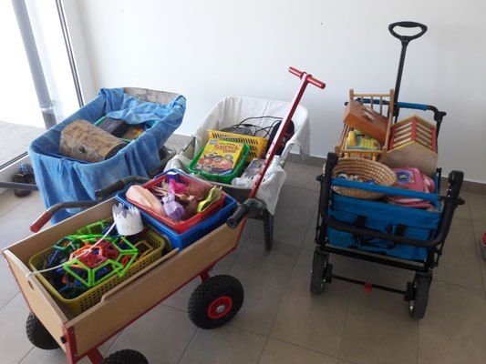 Mehrere Schubkarren voller Spielzeug, Bücher und anderer Gegenstände stehen auf einem gefliesten Boden. Ein Wagen ist mit bunten Plastikspielzeugen beladen, ein anderer mit Büchern und Spielzeug.