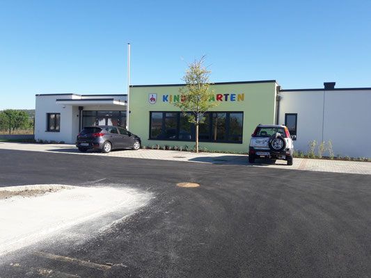 Ein Parkplatz vor einem grünen und weißen Gebäude mit der Aufschrift 'Kindergarten'. Zwei Autos sind davor geparkt.