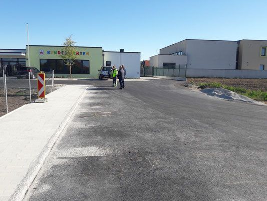 Drei Personen gehen vor einem Kindergartengebäude. In der Nähe ist ein Auto geparkt, und das Gebiet ist von einem Zaun umgeben.
