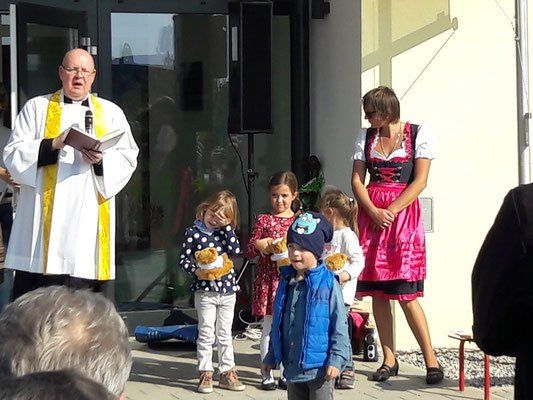 Ein Priester liest aus einem Buch, während eine Gruppe von Kindern und Erwachsenen Teddybären vor einem Gebäude halten.