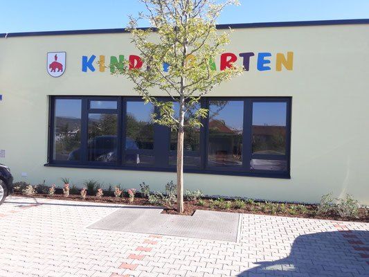 Die Vorderseite eines Gebäudes zeigt Kindergarten mit einem Logo darüber. Ein Baum steht davor, und es gibt Pflanzen darum herum. Ein Auto ist links geparkt.
