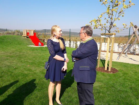 Ein Mann und eine Frau stehen auf einem grünen Rasen. Die Frau trägt ein Kleid und der Mann eine Brille. Hinter ihnen steht eine rote Rutsche und ein Baum ist auf der rechten Seite gepflanzt. Ein Zaun befindet sich hinter ihnen und einige Berge sind in der Ferne sichtbar.