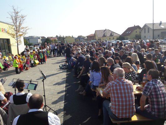 Viele Menschen, darunter Kinder in gelben Westen, versammeln sich im Freien in der Nähe von Tischen. Musikinstrumente sind auf der linken Seite aufgestellt, und Gebäude sind im Hintergrund.