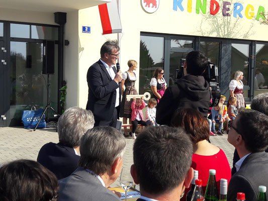 Ein Mann spricht in ein Mikrofon vor einer Menschenmenge und hält eine Flagge. Die Veranstaltung findet vor einem Gebäude mit der Aufschrift 'Kinderga' statt. Menschen sitzen und stehen, eine Kamera erfasst den Moment.