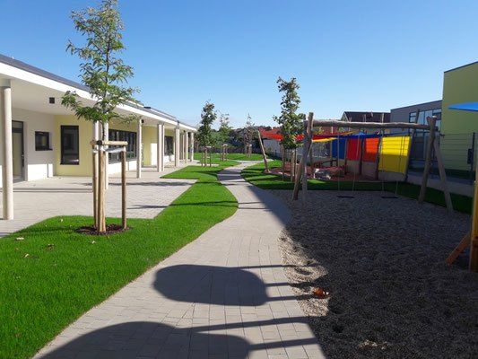 Ein Weg führt von einem modernen Gebäude zu einem Spielplatz mit bunten Rutschen und Schaukeln, umgeben von Bäumen und Gras unter einem klaren Himmel.