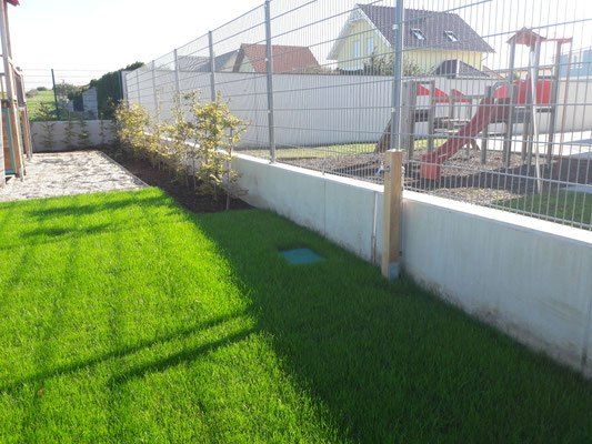 Ein Garten mit einer weißen Mauer, einem Metallzaun und einer roten Rutsche mit einem Spielbereich. Der Garten ist von Rasen und kleinen Bäumen umgeben.