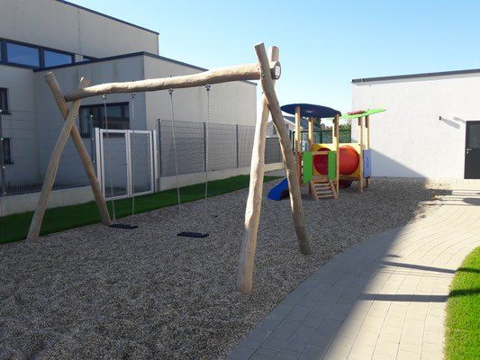 Ein Outdoor-Spielplatz mit einem Holzschaukelgestell, einer bunten Spielstruktur und einer Rutsche auf einer Schotterfläche.