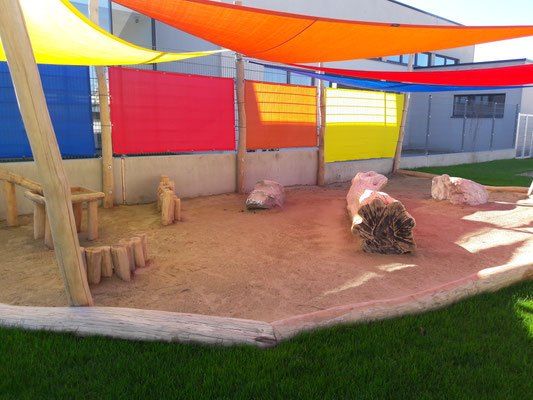 Ein bunter Outdoor-Spielplatz mit Holzbänken, Baumstämmen, Steinen und bunten Stoffzelten. Der Bereich ist von einer Steinmauer umgeben, im Hintergrund befindet sich ein Gebäude.