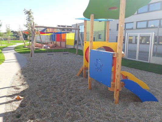 Ein Kinderspielplatz mit bunten Geräten, Schaukeln und einer Rutsche auf einer Kiesfläche, mit einem Gebäude und einem Zaun im Hintergrund.